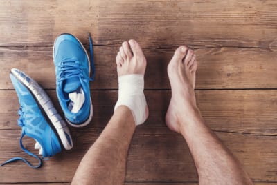 Recovery after running a marathon - Overhead shot of the bare legs of a white male with the left ankle bandaged and a pair of blue trainers next to the left foot on a wooden floor